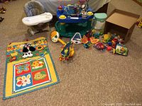 Wide view showing baby seat with tray, blue baby activity center/mobile, yellow floor blanket with plush dog and animal prints, and multiple small baby toys scattered
