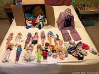 Wide view of Barbie dolls, small dolls, accessories, and clothing laid out on bed with box of clothing behind.