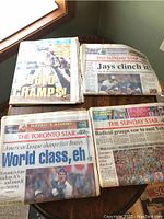 Four 1992 Toronto Blue Jays memorabilia newspapers stacked on a wooden surface near a window, blended headlines showing championship celebrations.