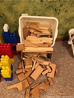 Plastic container filled with assorted natural wooden building blocks scattered on the floor next to large colorful interlocking plastic blocks arranged in towers.