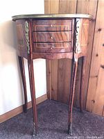 Full view of French style demi lune side table showing curved front, three drawers, brass trim along top edge and decorative metal accents on legs and sides