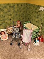 Photo showing two doll cribs, two doll strollers, and two dolls in front of patterned green wall, on carpeted floor.