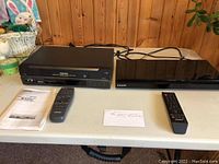 Photo showing Toshiba VCR Player model W-522C on left and Samsung Blu-ray Disc player on right with remotes and manual in front on table.