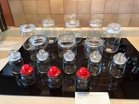 Front view of assorted glass storage containers of multiple sizes and shapes arranged on black surface, including jars with clear glass lids and some with red knob lids.