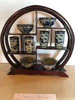 Full view of all 8 sake cups neatly arranged on circular wooden shelf, showing bowls and cylindrical cups with blue and green dragon motifs.