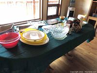 Full view of assorted kitchen items on green tablecloth including glass bowls, ceramic bowls, pitcher, and trays.