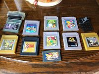 Photo showing all 11 Nintendo Game Boy game cartridges laid on a wood table, including GameShark, Pokémon, Zelda, Kirby, Mario, Yoshi, Jurassic Park, Dragon Ball Z, Balloon Kid, and a black accessory.