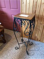 Full view of black metal plant stand with decorative scrollwork and wooden top, positioned on carpet near wooden wall paneling and pink door.