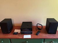 Sony black CD player with top-loading tray and buttons on front, sitting between left and right black Sony speakers with a remote control, external antenna, power cord, and instruction note laid out on wood surface.