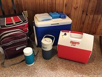 Photo showing all cooler items including a large soft-sided cooler bag, red and white Igloo Playmate Elite cooler, blue and white Coleman cooler, and two Thermos style water jugs with blue ice packs on top
