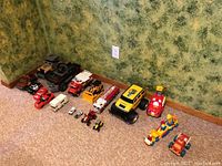 Lot of assorted vintage toy vehicles including trucks, cars, construction and emergency vehicles in various sizes and colors arranged on carpet in a room corner.