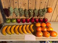 Display of various plastic decorative fruit including grapes, apples, pears, bananas, and oranges arranged on shelving with hanging strings visible.