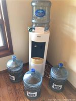 White Chicago refrigerated water cooler with black dispensing panel and two taps, top-mounted water container and 3 additional containers around base placed near wall corner.