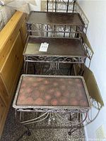 View of three rectangular nesting metal accent tables stacked inside each other, showing decorative metal scrollwork sides and dark tops with scratches and wear marks.
