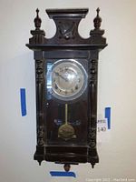 Full view of dark wood antique regulator clock with glass front, circular clock face and pendulum case showing distinct columns, ornate finials, and decorative carvings.