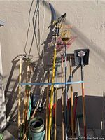 Photo showing long handled garden tools including tree saw, rakes, shovels, and picker tool leaning against a wall.