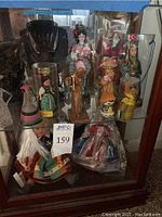 Full display cabinet photo showing multiple multicultural dolls, including Asian traditional dolls, wooden carved statues, and fabric costume dolls.