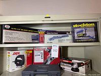 Shelf with boxed new power tools and tool sets including Skil jigsaw, Drill Master angle grinder, Craftsman auto-hammer, deep impact socket set, O-ring assortment, hex key set.