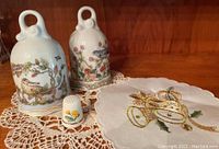 Photo shows two German porcelain bells labeled November and May, a small porcelain thimble, and embroidered lace doilies with detailed floral and horn designs.