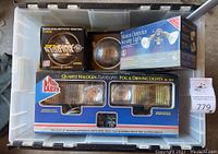 Plastic storage bin containing assorted automotive lights and parts, including boxed Night Blaster fog and driving lights, a round headlamp bulb, a box marked Borg-Warner automotive parts, and a motion detector security light boxed.