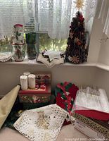 Photo of assorted holiday linens, a pinecone Christmas tree ornament, candles, boxes, and fabric pieces on a windowsill and table surface.
