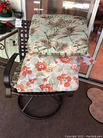 Outdoor metal mesh swivel chair with floral cushion on seat and two extra cushions stacked on top showing different floral prints and visible white paint spot on one cushion.