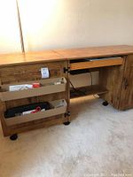 Wood sewing table with fold-down left side, drawer and door open showing storage bins with sewing supplies including scissors, pins, and thread.