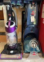 Front view of both vacuum and carpet cleaner shows machines side by side; vacuum is purple and black with clear catch canister, carpet cleaner is blue with control panel.