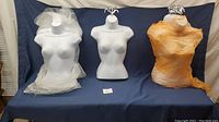 Three female half mannequins, two wrapped in plastic, one unwrapped, displayed on blue fabric. The middle one is unwrapped and shows full front torso.