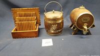 Front view of three items: wooden box with dividers, oak barrel biscuit container with metal handle and lid, and oak barrel dispenser with spigot on wooden stand.