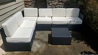 Arranged L-shaped configuration of six dark gray wicker resin seating modules with white cushions and square coffee table on concrete surface