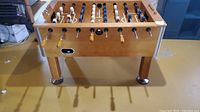 Front angle of the Harvard foosball table showing wooden frame, metal rods with wooden handles, and part of the playing field with blue and white player figures.