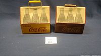 Both wooden Coca-Cola bottle carriers together showing front and side views, with cloth covers displaying three raised bottle images each and yellow-handled metal carrying grips.