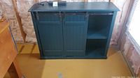 Front view of dark teal painted TV stand with barn sliding doors closed and one open shelf visible on right side. Cabinet top holds attached power strip. Basement room background with insulation on walls.