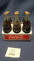Front view of aluminum Coca Cola six-pack carrier with six embossed glass bottles filled with dark liquid and rusty caps.