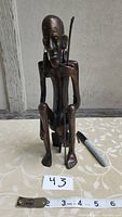 Full front view of copper man figurine sitting with staff, placed next to ruler showing height about 11 inches and width about 4 inches.