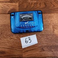 Front view of the blue sparkle Fender Automatic Guitar Tuner showing the analog meter and Fender logo on wooden surface with numbered lot card.