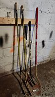 Five hockey sticks standing against a stained concrete wall in a basement or storage area, showing full length and condition.