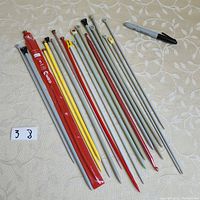 Photo showing the full set of 20 knitting needles in assorted sizes and colors laid out on a light patterned cloth, including a black marker for scale.