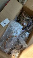 Photo showing a cardboard box filled with multiple pairs of prescription glasses in assorted frames, mostly wire and plastic, in various colors and shapes, some stored in clear plastic bags.