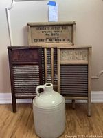 Three antique wooden washboards with metal scrubbing surfaces and a ceramic jug in front on wooden floor, showcasing all items together.