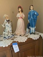 Three porcelain figurines on a wooden cabinet with a lace runner, including a lady with a dog and beads, a young woman in pink, and a man in blue period attire.