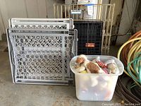 Two plastic pet gates and black metal dog crate stacked against a wall with a plastic bin of dog toys next to them.