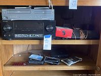 Magnavox DVD/VHS combo player on top shelf with front buttons and disc tray visible. Below is Pioneer stereo system with dual large knobs on front, black finish, control buttons and display panel. To the side is boxed Microsoft LifeCam HD-3000 and wired items.