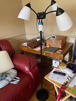 Full view of a black metal floor lamp with three white plastic shades, standing beside a red chair and wooden table.