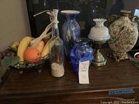 Overall view of wooden surface displaying a variety of glass and ceramic decorative items including faux fruit bowl, glass bottles and vases, ceramic vases, and lamp