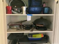 Shelf showing various kitchen items including two stovetop espresso makers, a black griddle with glass lid, blue mixing bowls, and a wok with wooden handle and lid.