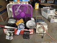 Photo showing various fans, work lights, flashlights, a headlamp in box, and a small radio on garage floor
