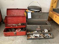 Photo showing one red metal toolbox filled with grinding discs and hand tools, two gray metal tool boxes with tops open revealing assorted hardware and hand tools, and a black and yellow tool bucket behind.