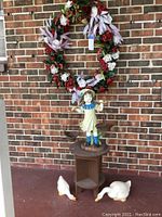 Front view of the entire lot including the wreath hanging on brick wall, statue of girl on wood plant stand, and two ceramic geese on porch floor.